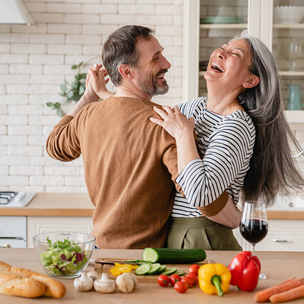Couple dancing and laughing together in a kitchen with fresh vegetables and wine.
